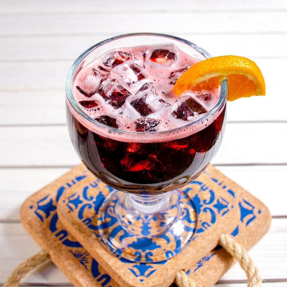 Red sangria with ice and an orange slice, on cork coasters with blue designs.