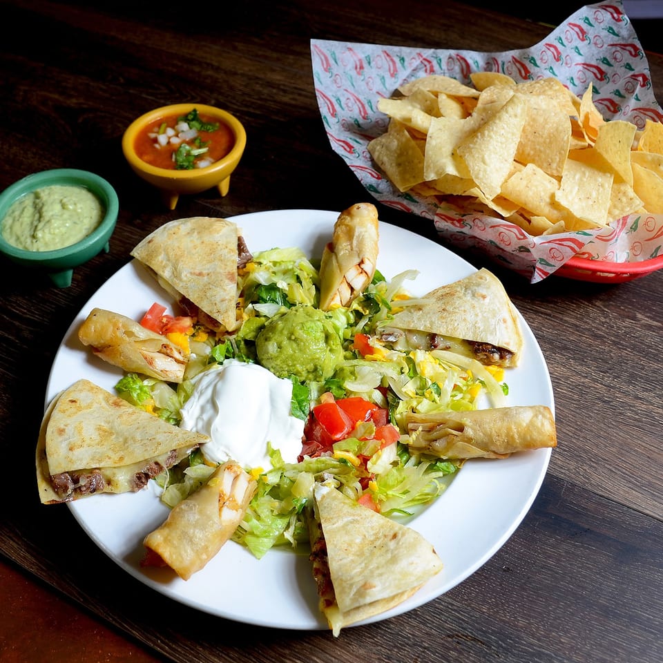 A platter of quesadillas, taquitos, guacamole, sour cream, and salsa, with a basket of chips.