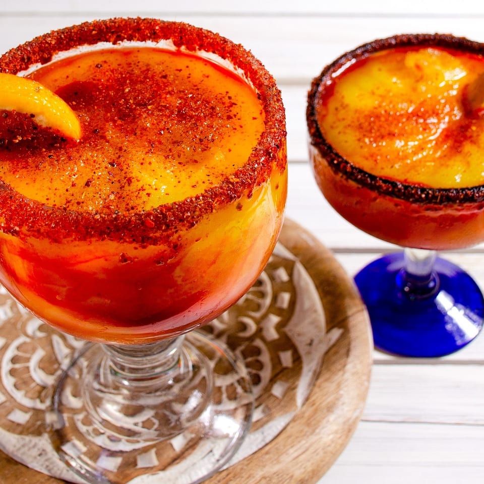 Two vibrant orange and red frozen drinks with chili-lime rims and orange slices on a white wooden table.