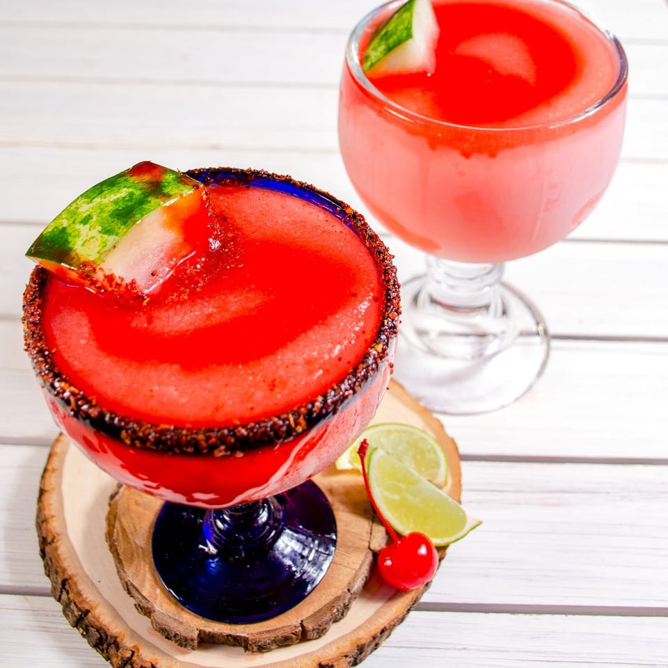 Two red frozen drinks, one garnished with watermelon, lime, and a cherry on a wooden coaster.