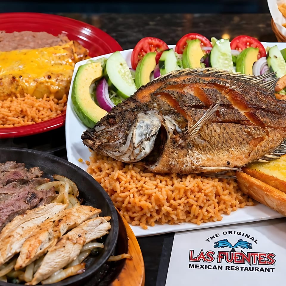 A table laden with various Mexican dishes, including whole fried fish, fajitas, enchiladas, and rice.