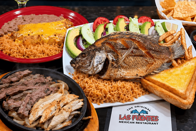 A table laden with various Mexican dishes, including whole fried fish, fajitas, enchiladas, and rice.
