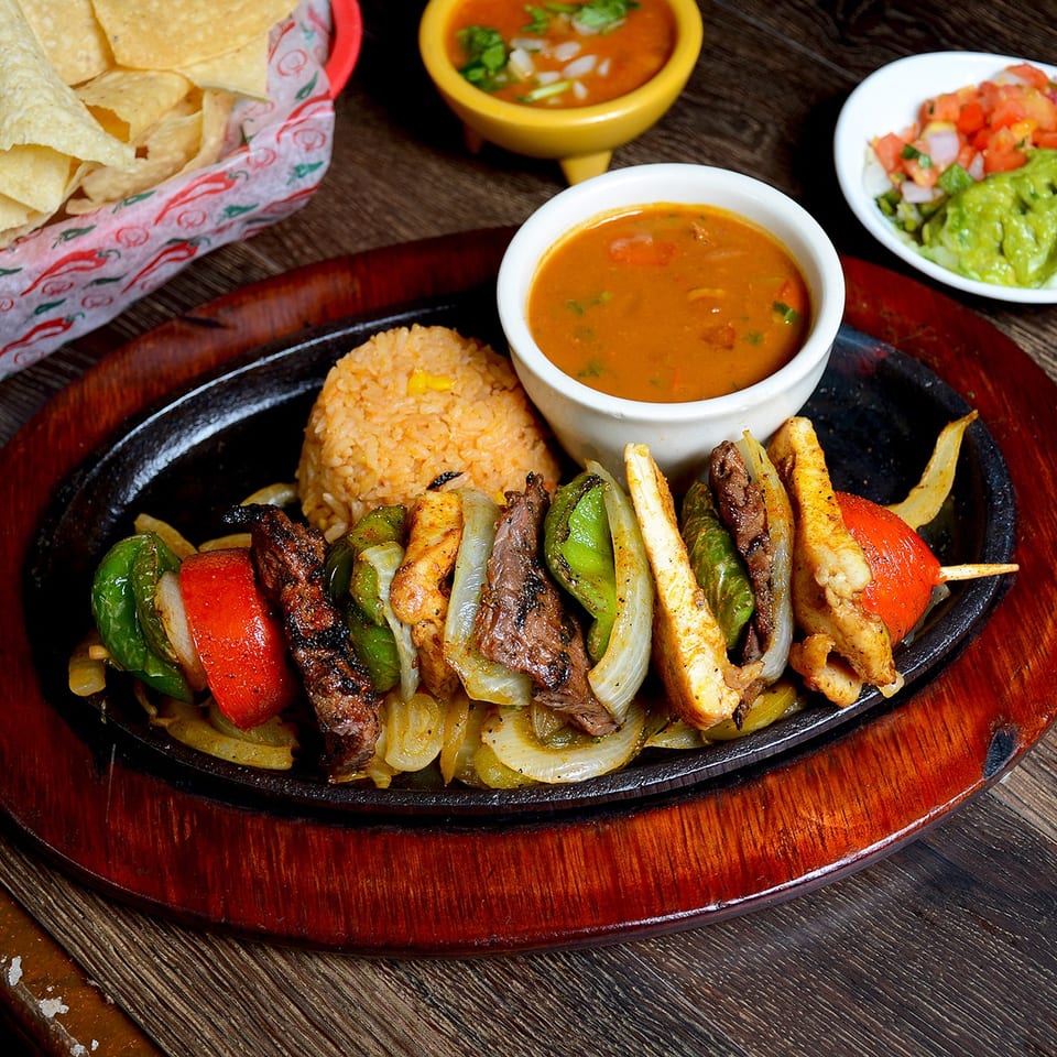 Sizzling fajita skewers with steak, chicken, and vegetables, served with rice, beans, chips, and guacamole.