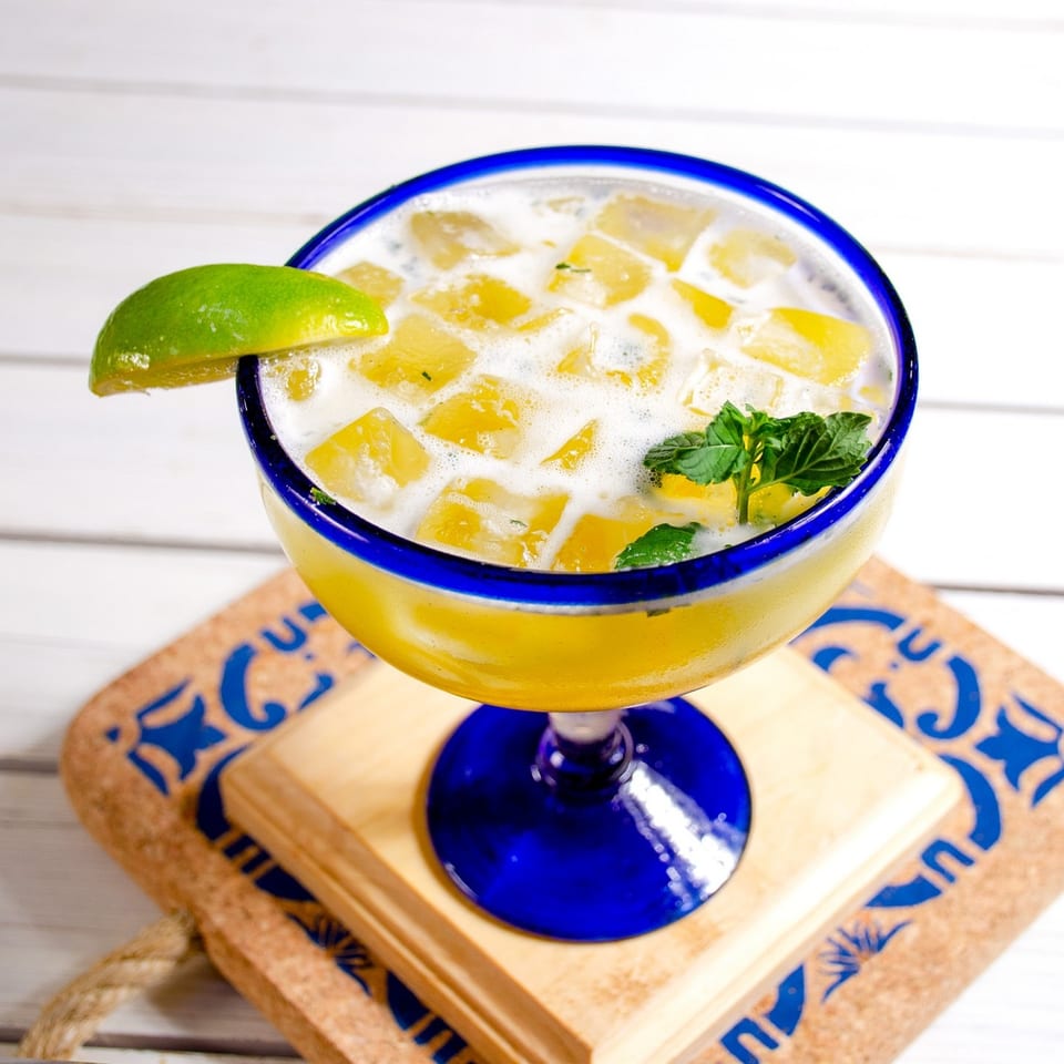 A vibrant yellow drink with ice, lime, and mint in a blue-rimmed glass on a patterned coaster.